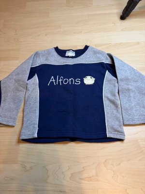 Blå och grå Alfons Åberg tröja - Mysig långärmad tröja i blått och grått med Alfons Åberg-tryck och texten 'Alfons' på bröstet. Tröjan har rund halsringning och kontrastfärgade ärmar. Perfekt för dig som gillar klassiska barnmotiv.