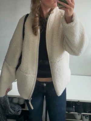 Krämvit teddyjacka med dragkedja - Mysig krämvit teddyjacka med oversized passform och dragkedja framtill. Jackan har stora fickor på sidorna och är gjord i fluffigt teddy-material som ger en trendig look. Perfekt för att hålla värmen under kyliga dagar och passar till jeans eller kjol.