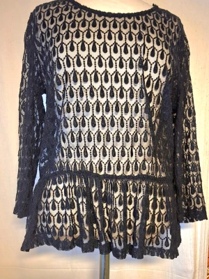 Svart spetsblus med volang - Snygg svart spetsblus med unikt droppformat mönster och volang nedtill. Blusen har rund halsringning och långa ärmar. Materialet är genombruten spets som ger en cool och trendig look. Perfekt att styla över ett linne eller topp.