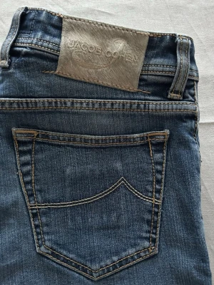 Jacob Cohen 622/Nick - Säljer ett par klassiska blå jeans från Jacob Cohen med raka ben och snygga slitningar i storlek W31. Jeansen har fem fickor, kontrastsömmar och en exklusiv patch i mocka baktill. Tillverkade i mjuk denim med skön stretch för extra komfort. Reparerat hål under dragkedjan och i vänstre fickan, båda osynliga vid användning och fungerar som nya.