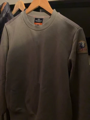 Grön sweatshirt från Parajumpers - Snygg grön sweatshirt från Parajumpers med rund halsringning och ribbade muddar. På vänster ärm sitter ett tydligt Parajumpers-märke med broderad logga och texten 'PJS Anchorage'. Perfekt för en clean och avslappnad stil. Köpt på Johnells, knappt använd!!!