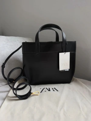 Svart handväska från Zara - Stilren svart handväska från Zara i slätt konstläder. Väskan har både handtag och avtagbar axelrem i samma material. Invändigt finns ett fack med dragkedja och magnetknäppning. Perfekt för dig som gillar minimalistisk design.