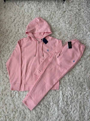 Rosa loungewear-set från Polo Ralph Lauren - Mysigt loungewear-set i ljusrosa från Polo Ralph Lauren. Setet består av en hoodie med dragkedja och ficka framtill samt matchande mjukisbyxor med resår i midjan. Båda plaggen har den klassiska blå Polo-loggan broderad. Perfekt för chill dagar hemma.