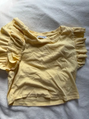 Gul ribbad topp med volangärm från Zara - Supersöt ljusgul ribbad topp från Zara med volangärmar som ger en extra touch till outfiten. Croppad modell med rund halsringning och mjukt stretchigt material. I storlek M.💛