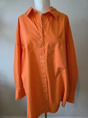 H&M Oversized orange skjorta i bomull st M - Snygg oversized skjorta i klar orange färg med klassisk krage och bröstficka från H&M/Divided Skjortan har långa ärmar och knappar framtill. Tillverkad i mjuk bomull som känns skön mot huden. Perfekt för dig som gillar en avslappnad och trendig stil.