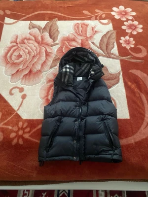 Burberry Väst/jacka - Snygg svart Burberry Väst/jacka med avtagbar huva och dragkedja framtill. Västen har två stora fickor med dragkedja och ett rutigt innerfoder i huvan. Perfekt för lager-på-lager och streetwear-stil.