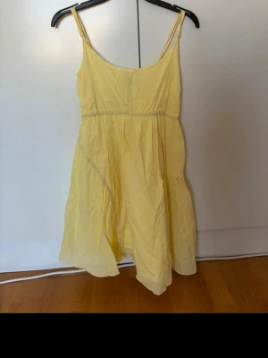 Gul sommarklänning med smala axelband - Söt gul klänning med smala axelband och utställd kjol. Klänningen har en enkel, luftig design och är perfekt för varma dagar. Materialet känns lätt och bekvämt, och modellen är kort med en avslappnad passform.