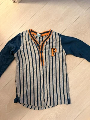 Randig henley från Polarn O. Pyret - Snygg henley från Polarn O. Pyret med blå och grå vertikala ränder, orange detaljer och bokstaven P på bröstet. Långärmad modell med knappar vid halsen. Perfekt för en sportig och avslappnad stil.