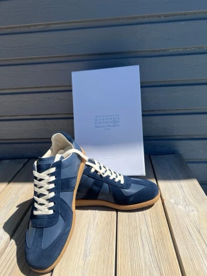 Blå Maison Margiela sneakers - Snygga blå sneakers från Maison Margiela med vita skosnören och gummisula. Skorna har en mix av mocka och textilmaterial i olika blå nyanser och klassisk låg siluett. Perfekta för dig som gillar stilrena och trendiga sneakers.