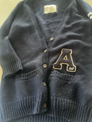 Mörkblå kofta från Abercrombie & Fitch - Mysig mörkblå kofta med knappar framtill och två fickor. Stor broderad bokstav 'A' med texten 'A&F' på vänster sida. Klassisk collegestil med ribbade muddar och v-ringning. Perfekt för en avslappnad och trendig look.