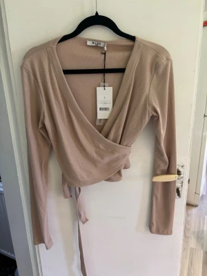 Beige omlottopp från NA-KD - Trendig beige omlottopp från NA-KD i ribbad bomullsmix. Toppen har långa ärmar, djup v-ringning och knytband i midjan för en snygg lätt croppad passform. Perfekt att styla med jeans eller kjol för en cool och avslappnad look.