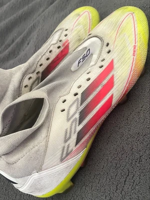 Adidas F50 vita fotbollsskor - Säljer ett par Adidas F50 fotbollsskor i vitt med neon-gula och röda detaljer. Skorna har klassiska tre ränder på sidan och en strömlinjeformad design utan snörning. Ovandelen är i syntetmaterial för lätt vikt och bra bollkontroll. Perfekta för snabba spelare på planen.