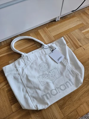 Isabel Marant Tote Bag Utsmyckad - Snygg vit shoppingväska från Isabel Marant i kraftigt tyg. Väskan har dubbla handtag och är dekorerad med silverfärgade nitar som bildar Marant-loggan och texten på framsidan. Perfekt för att bära allt du behöver till skolan, stranden eller stan.