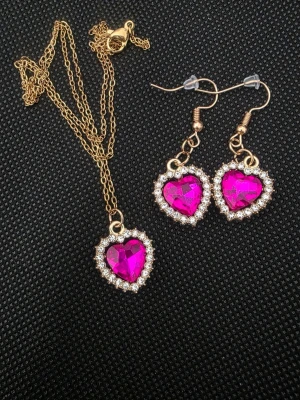 Set med hjärtformad bodykedja och örhängen - Snyggt handgjort set med guldfärgad bodykedja och matchande örhängen, båda med hjärtformade rosa stenar omgivna av glittrande små kristaller. Perfekt för att ge din outfit extra bling och romantisk touch.