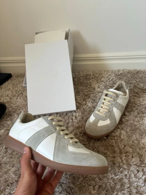 Maison Margiela sneakers i vitt och grått - Maison Margiela sneakers med klassisk låg profil, vit läderbas och ljusgrå mockadetaljer. Beigea snören och bruna gummisulor ger en clean och stilren look. Perfekt för dig som vill ha något exklusivt och trendigt.