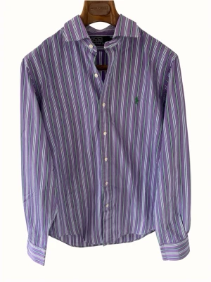 Lila randig skjorta från Polo Ralph Lauren - Klassisk lila skjorta från Polo Ralph Lauren med tunna vita och gröna ränder. Skjortan har lång ärm, knappar framtill och en liten grön broderad logga på bröstet. Tillverkad i mjuk bomull och perfekt för en stilren look.