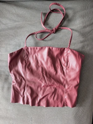 Vinröd croppad linne-topp i skinnimitation - Säljer en vinröd croppad linne-topp med tunna axelband och knyt i nacken. Toppen är gjord i skinnimitation och har en enkel, stilren design utan mönster. Perfekt för dig som vill ha en edgy och trendig look.