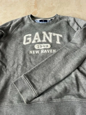 Grå sweatshirt från GANT - Klassisk grå sweatshirt från GANT med tryck 'GANT 1949 NEW HAVEN' i vitt framtill. Tröjan har rund halsringning, långa ärmar och ribbade muddar vid ärmslut och nederkant. Perfekt för en avslappnad och sportig stil.