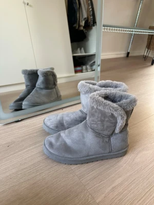 Gråa fluffiga tofflor i mocka - Mysiga gråa tofflor med mjukt, fluffigt foder och ovansida i mocka. De har en rund tå och mellanhögt skaft som håller fötterna varma. 
