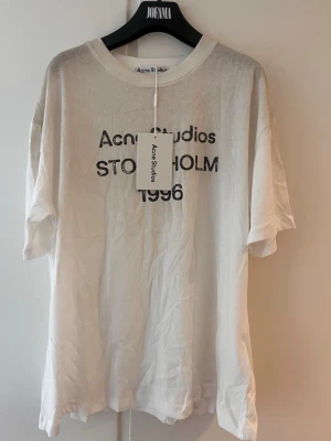 Vit Acne Studios t-shirt med tryck - Vit t-shirt från Acne Studios med svart texttryck på bröstet där det står 'Acne Studios Stockholm 1996'. Klassisk rund hals och korta ärmar. Tillverkad i mjuk bomull för en clean och stilren look.