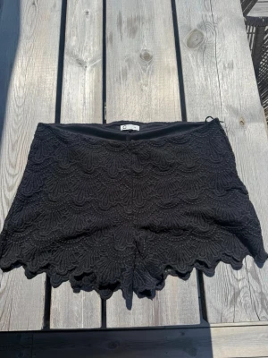 Svarta spets-shorts med volangkant - Säljer ett par svarta shorts från cubus med snygg spets och volangkant nertill. De har en feminin look med detaljerat mönster i spetsen och sitter skönt i midjan. Perfekta för varma dagar när du vill ha något både stilrent och trendigt.