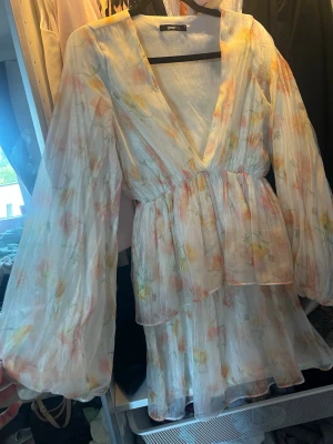 Blommig kortklänning med volang ginatricot - Söt kortklänning från Sisters Point i ett tunt, luftigt material med blommigt mönster i pastellfärger. Klänningen har v-ringning, volangdetaljer och långa, vida ärmar. Perfekt för sommarens alla tillfällen!