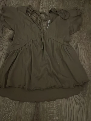 Brun kortärmad blus med knytband - Snygg brun blus med korta ärmar och v-ringning med knytband i halsen. Blusen har en lös passform och volangkant nedtill. Perfekt för en avslappnad och trendig look. Materialet känns lätt och luftigt.