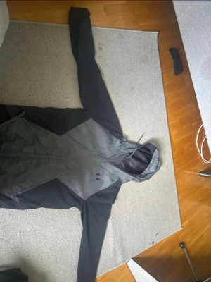 Svart vindjacka från Under Armour - Snygg svart vindjacka från Under Armour med dragkedja framtill och huva. Jackan har en sportig look med två sidofickor och är tillverkad i ett lätt, vindtätt material. Perfekt för blåsiga dagar och outdoor-aktiviteter.