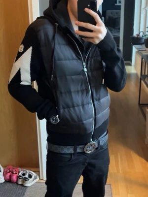 Svart cardiganjacka från Moncler - Svart cardiganjacka från Moncler med vadderad kropp och stickade ärmar. Jackan har en stor vit zigzagdetalj på ena ärmen och Moncler-logga. Utrustad med huva och dragkedja framtill. Materialmix av quiltad nylon och mjuk stickad bomull för en modern look.