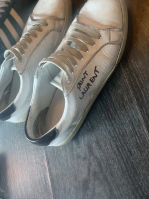 Vita Saint Laurent låga sneakers - Säljer ett par vita låga sneakers från Saint Laurent med svart detalj på hälen och klassisk snörning. Skorna har en stilren design med Saint Laurent-logga på sidan och är tillverkade i läder.