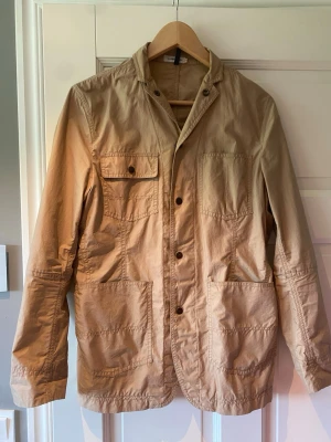 Beige overshirt från J.Lindeberg - Stilren beige overshirt med flera stora fickor framtill och knappstängning. Overshirten har en avslappnad passform, klassisk krage och är tillverkad i ett lätt bomullstyg. Perfekt lager på lager-plagg för en casual look.