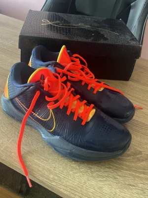 Nike Kobe 5 Protro caitlin Clark PE - Säljer ett par Nike Kobe 5 Protro basketskor i mörkblått med orange och gula detaljer. Skorna har en låg profil, ventilerande mesh på tungan och starka orangea skosnören. Perfekta för dig som vill sticka ut på basketplanen.