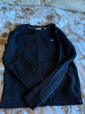 Svart sweatshirt med liten logga - Svart sweatshirt med rund hals och liten broderad logga på bröstet. Tröjan har långa ärmar och ribbade muddar vid ärmslut och nederkant. Perfekt för en avslappnad och stilren look.