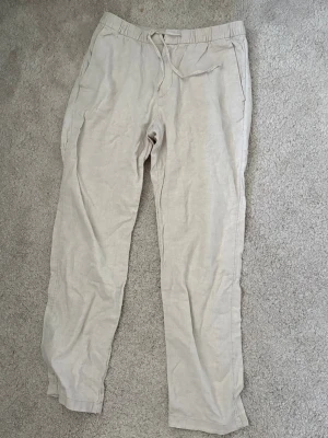 Beiga linnebyxor - Snygga och avslappnade beiga linnebyxor med elastisk midja och dragsko. Byxorna har raka ben, sidfickor och en bakficka. Perfekta för en chill och stilren look. Materialet är lätt och luftigt linne.
