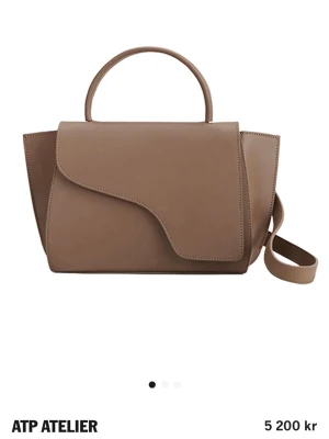 Hazelnut  handväska från ATP Atelier - Säljer en snygg beige handväska från ATP Atelier i slätt läder. Väskan har ett unikt lock med asymmetrisk form, ett topphandtag och en avtagbar axelrem. Perfekt för dig som gillar stilrena och moderna accessoarer. Skriv för fler bilder. små repor 