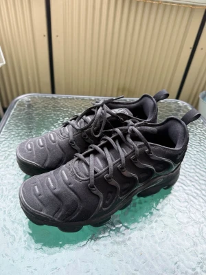 Vapormax svarta helt nya nästan  - Säljer ett par helsvarta Nike Air Vapormax Plus sneakers med chunky sula och ikoniska vågformade detaljer på ovandelen. Skorna har textil och syntetmaterial, snörning och dragflik bak. Perfekta för dig som gillar sportig och modern stil.