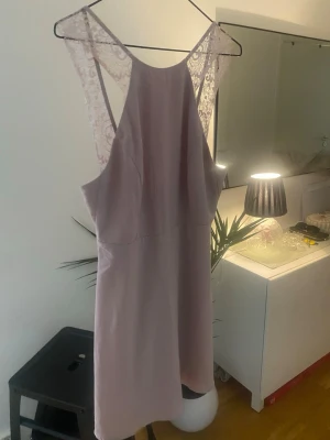 Ljuslila klänning med spetsdetaljer - Säljer en ljuslila kortklänning med vackra spetsdetaljer på ryggen och axelbanden. Klänningen har en ärmlös design och en enkel, elegant siluett som ger en feminin look. Perfekt för sommarens alla tillfällen!