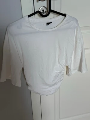 Vit topp med rynkade detaljer - Säljer en vit topp med rynkade detaljer på sidan och vid ärmslut. Toppen har rund halsringning och halvlånga ärmar. Perfekt att styla med jeans eller kjol för en clean look.
