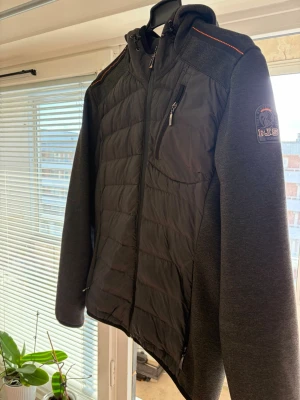 Svart pufferjacka från Parajumpers - Snygg svart pufferjacka från Parajumpers med quiltad front, mjuka stickade ärmar och huva. Jackan har dragkedja framtill, bröstficka med zip och orange detalj på axeln. Märkespatch på ärmen ger extra stilpoäng. Perfekt för kalla dagar.