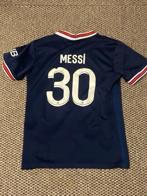 PSG matchtröja Messi #30 blå - Snygg mörkblå PSG matchtröja från Nike med Messi och nummer 30 på ryggen. Tröjan har vita och röda detaljer på krage och ärmslut, samt PSG-logga och sponsortryck på bröstet. Perfekt för dig som älskar fotboll och vill stötta Paris Saint-Germain.