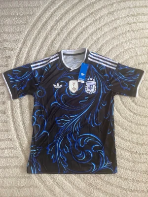 Argentina Adidas matchtröja svart/blå - Snygg Argentina matchtröja från Adidas i svart med blått och turkost mönster. Tröjan har korta ärmar, vita detaljer och officiella AFA- och FIFA-märken på bröstet. Tillverkad i lätt och funktionellt material, perfekt för fotboll eller träning.