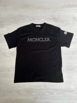 Svart Moncler t-shirt med logga - Snygg svart t-shirt från Moncler med stor vit logga på bröstet och Moncler-märke på ärmen. Klassisk rund halsringning och rak passform. Tillverkad i mjuk bomull som känns skön mot huden. Perfekt för dig som gillar stilrena plagg med exklusiv känsla.