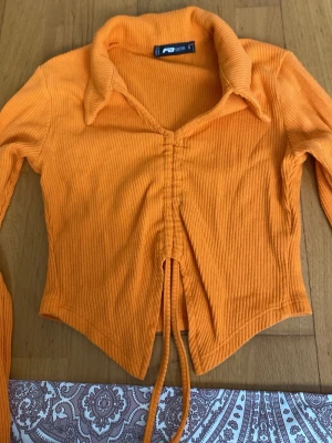 Orange ribbad croppad topp från Sister - Trendig orange croppad topp från Sister med långa ärmar och krage. Toppen är ribbad och har snörning framtill som ger en snygg omlott-effekt. Perfekt för dig som vill sticka ut med färg och coola detaljer.
