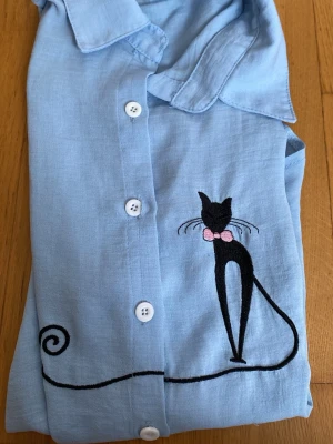 Ljusblå skjorta med kattbroderi - Unik ljusblå skjorta med korta ärmar och knappar framtill. På bröstet finns ett broderi av en svart katt med rosa rosett och lång svans. Skjortan har klassisk krage och är tillverkad i ett tunt, mjukt tyg som känns svalt och skönt.
