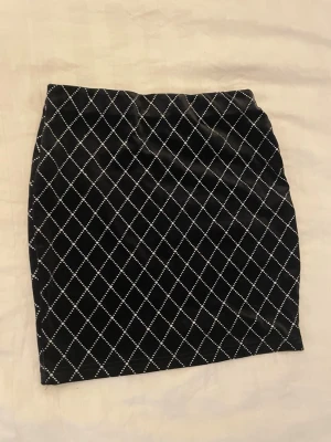 Svart kortkjol med rutigt mönster - Snygg svart kortkjol med vitt rutigt mönster. Kjolen har en rak passform och är perfekt för dig som gillar en stilren men ändå trendig look. Materialet känns mjukt och följsamt mot kroppen.