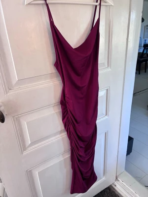 Vinröd långklänning med drapering - Vinröd långklänning med smala axelband och snygg drapering längs sidan. Klänningen har en figurnära passform och är tillverkad i ett mjukt, stretchigt material som ger en elegant siluett. Perfekt för dig som vill sticka ut med en stilren look.