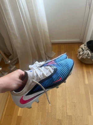 Nike phantom - Säljer ett par Nike fotbollsskor med blå ovandel och vita partier, samt svarta och rosa detaljer. Skorna har snörning och en låg profil, perfekt för snabba rörelser på planen. Swoosh-loggan syns tydligt på sidan och hälen. Materialet är syntetiskt för bra grepp och komfort.