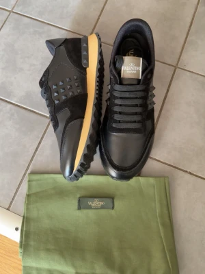 Svarta Valentino sneakers med nitar - Snygga svarta sneakers från Valentino med coola pyramidformade nitar på sidorna. Skorna har en beige sula med svarta detaljer och är gjorda i en mix av läder och textil. Klassisk snörning och logga på plösen. Perfekta för dig som vill sticka ut med stil.