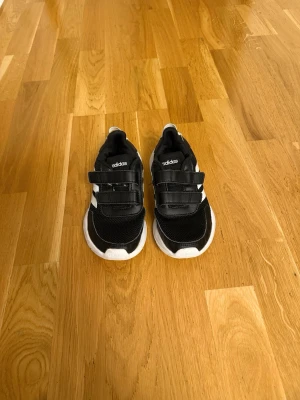 Svarta Adidas sneakers med kardborre - Snygga svarta sneakers från Adidas med vita detaljer och kardborreband för enkel påtagning. Skorna har en vit sula med greppvänligt mönster och klassiska tre ränder på sidan. Ovandelen är i mesh och syntetmaterial för bra andningsförmåga.
