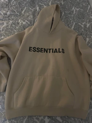 Beige Essentials Fear of God hoodie - Säljer en beige hoodie från Essentials Fear of God med svart tryck på bröstet. Hoodien har stor huva, känguruficka och ribbade muddar. Tillverkad i mjukt bomullsmaterial som är perfekt för en avslappnad streetwear-look.